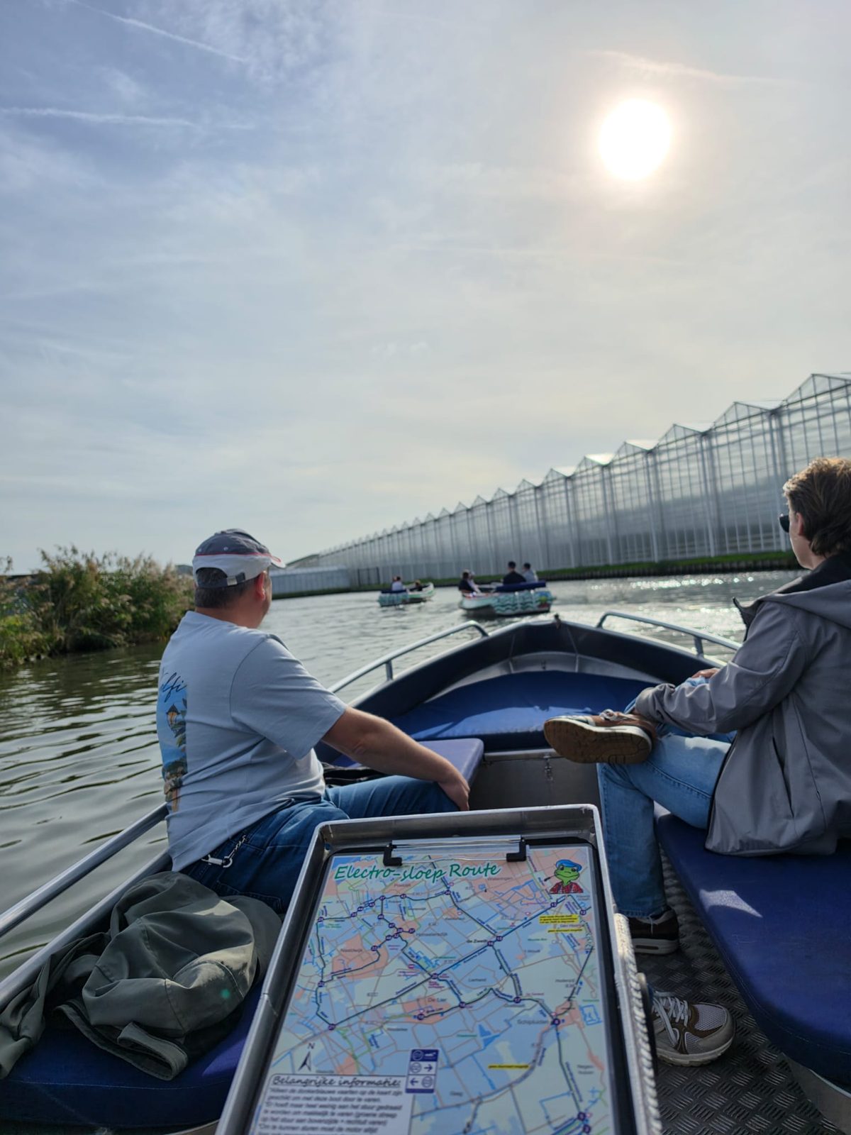 Twee collega's in een elektrische sloep met de routekaart van de Electro-sloep Route voorop, met kassen langs het water en twee andere bootjes voor ons