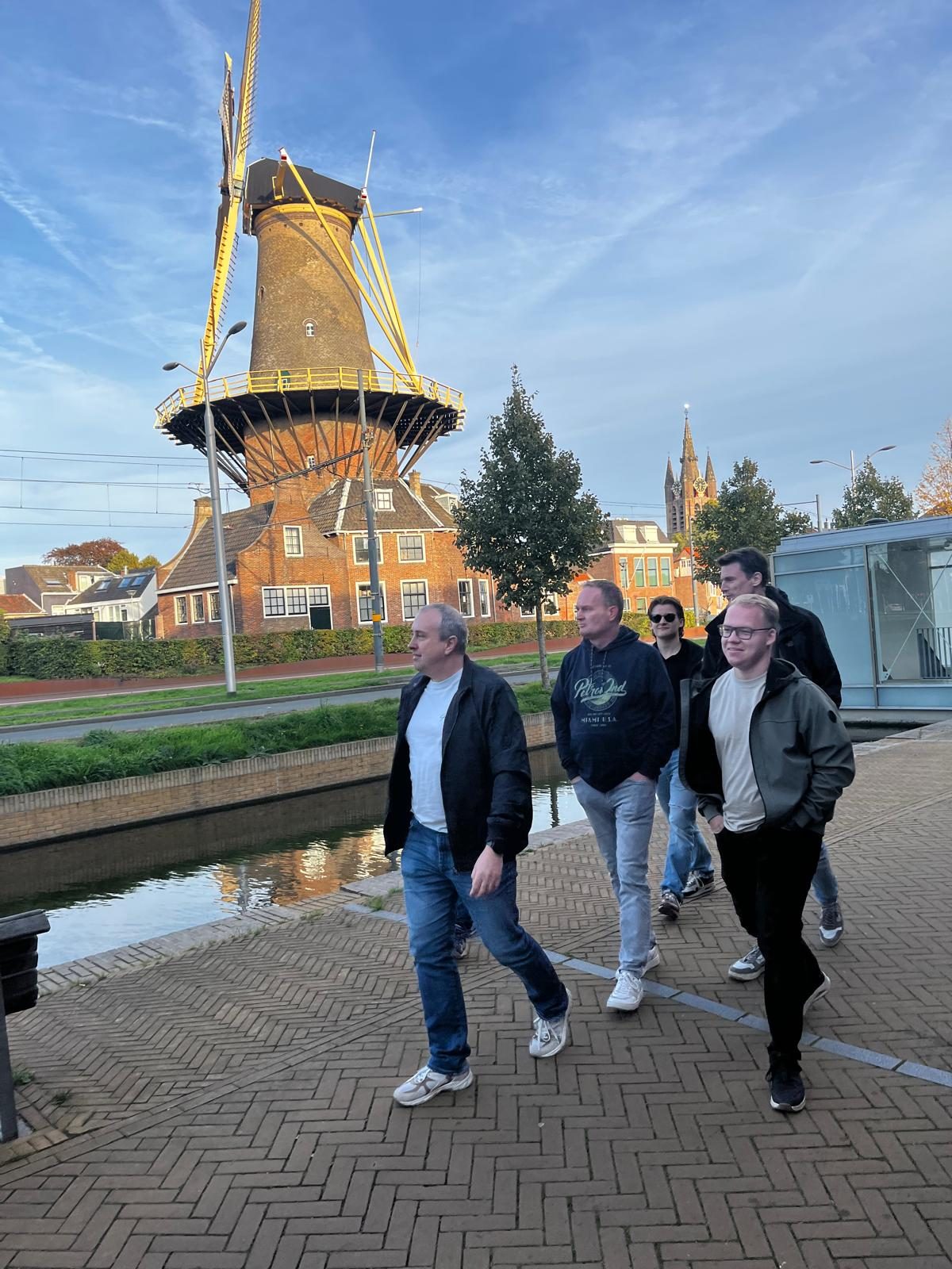 Vijf collega's wandelen langs een gracht in Delft, met een molen en een kerktoren op de achtergrond